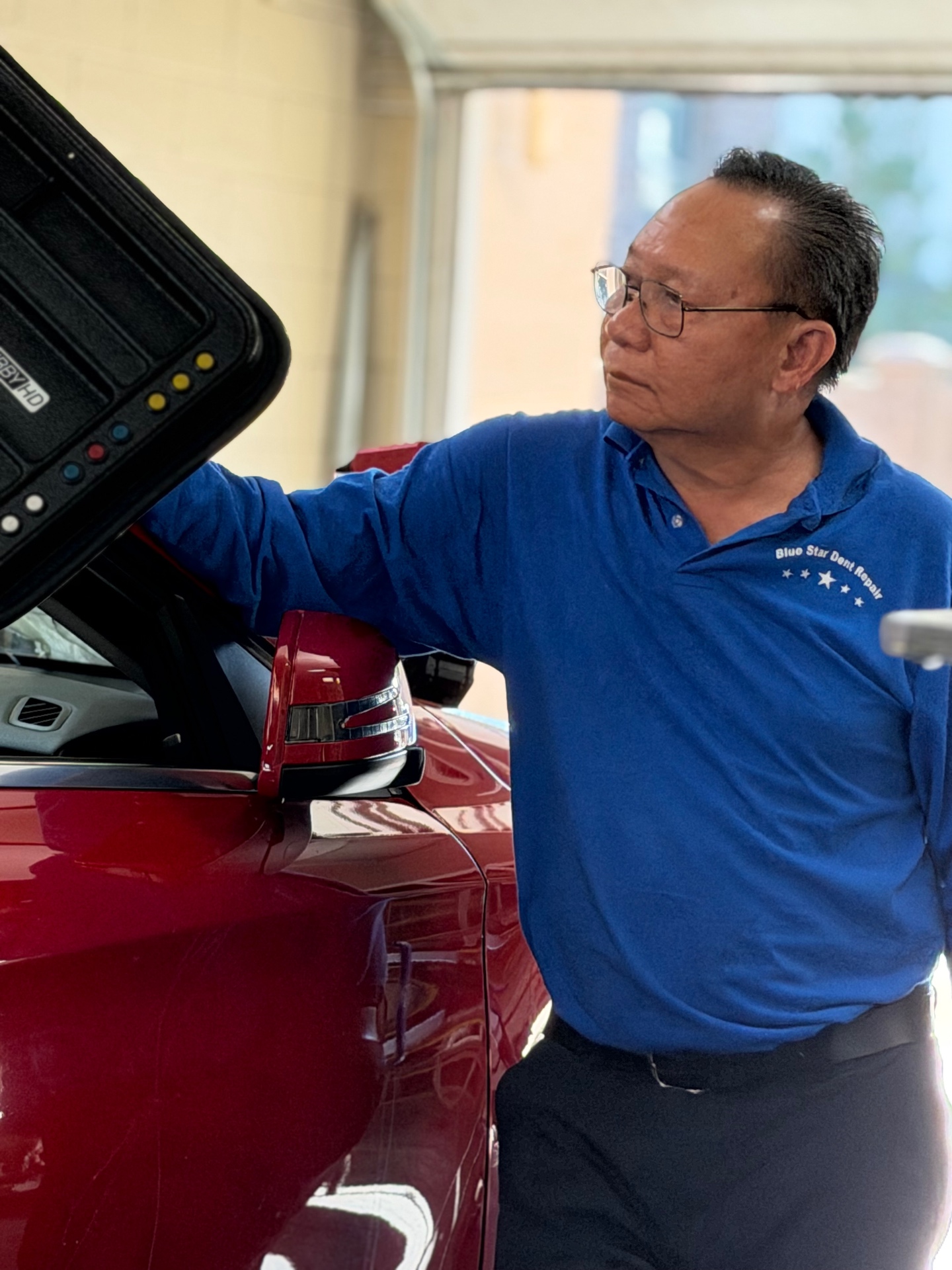 Technician inspecting a red vehicle panel beside a paintless dent repair light board